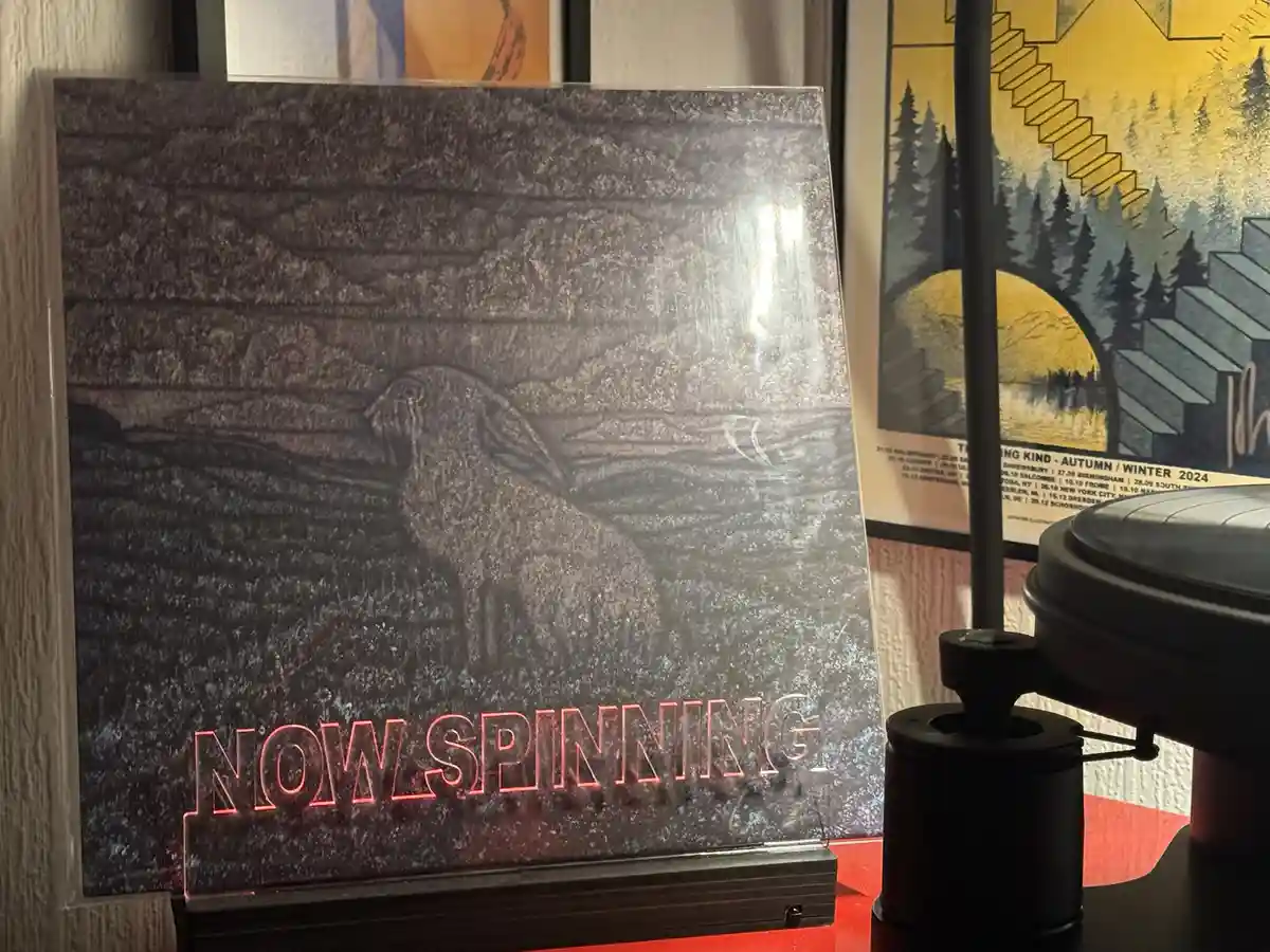 A vinyl record sleeve featuring a textured, dark gray illustration of a sheep-like creature standing in a field, with…