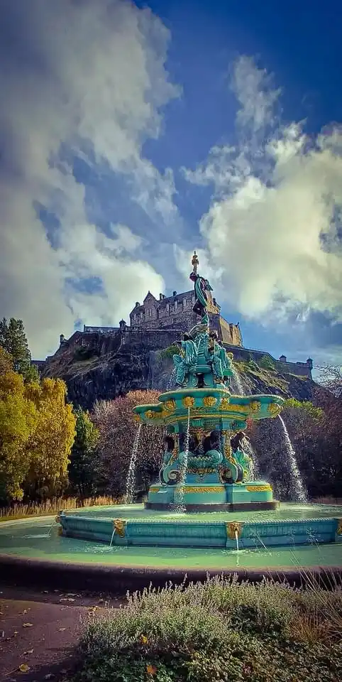 A vibrant, ornate teal-colored fountain with multiple tiers and water jets is centered in the foreground, set against a…