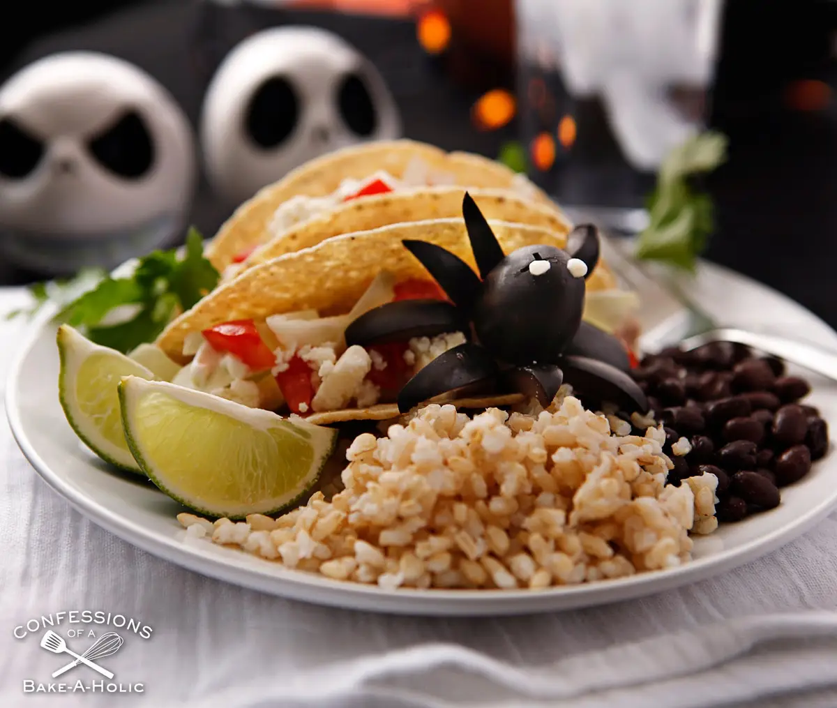 A plate of tacos, brown rice, and black beans is garnished with lime wedges and a decorative black olive shaped like a bat…