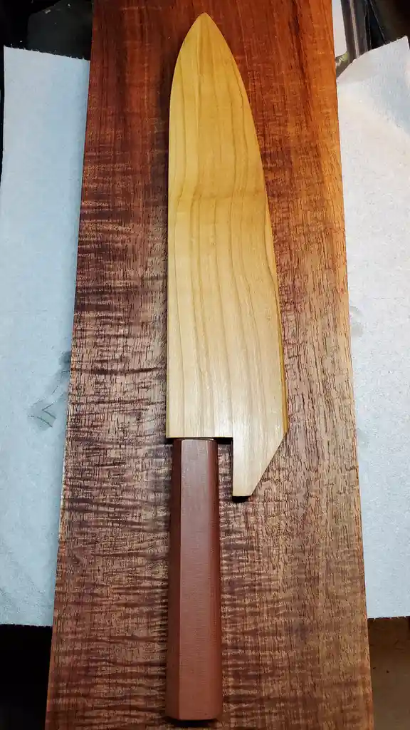 A wooden object shaped like a kitchen knife with a light-colored blade and dark brown handle rests on a dark wood surface…