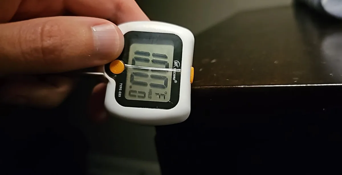 A hand holds a white digital thermometer with a probe inserted into a dark surface, displaying the reading '88.88' in large…
