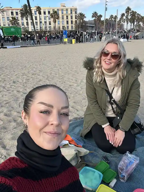 Two women are taking a selfie on a sandy beach with buildings and palm trees in the background. The woman in the foreground…