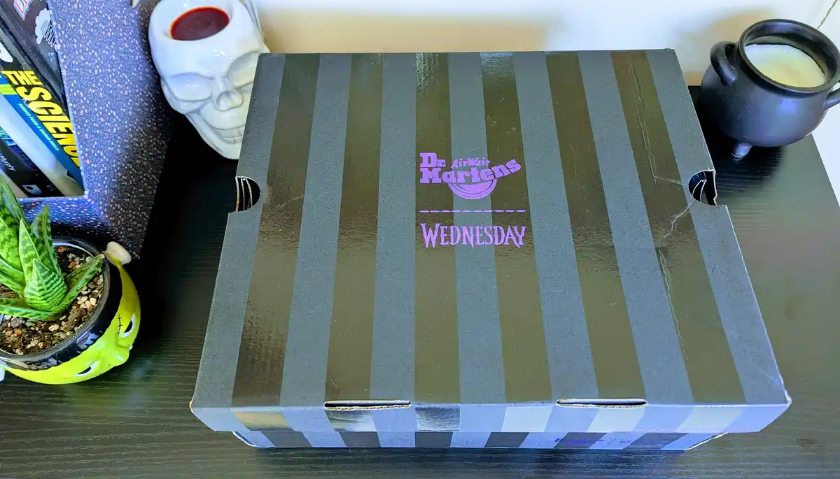 A Dr. Martens shoebox with black and olive green vertical stripes is shown on a dark wooden surface. The box has purple text…