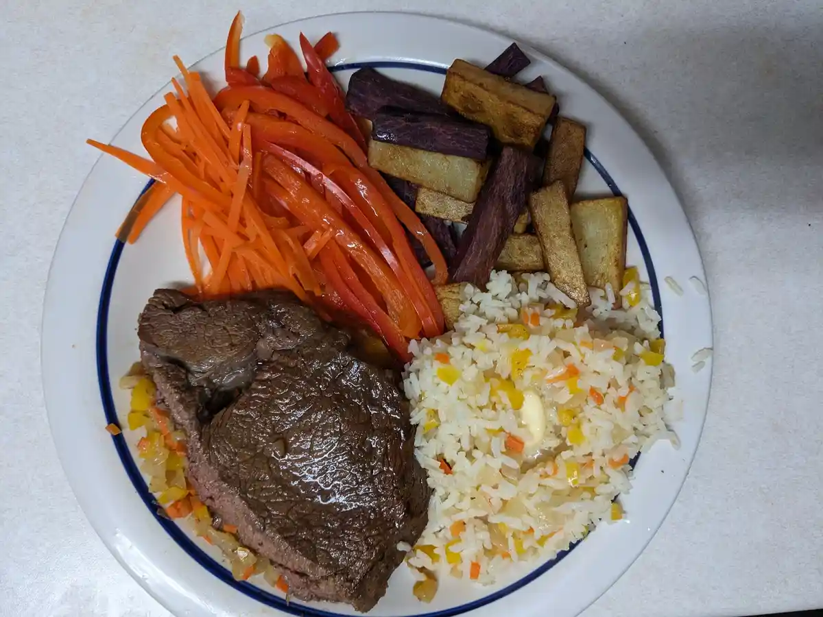 A white plate with a blue rim holds a meal consisting of a large piece of cooked beef, a serving of mixed rice with…