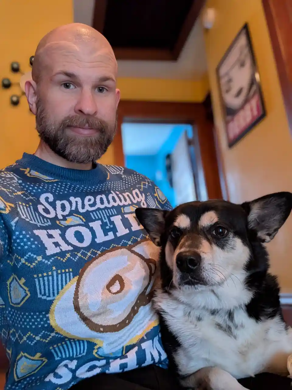 A bald man with a beard wearing a blue holiday sweater that says 'Spreading HOLIDAY CHEER' while holding a black and white…