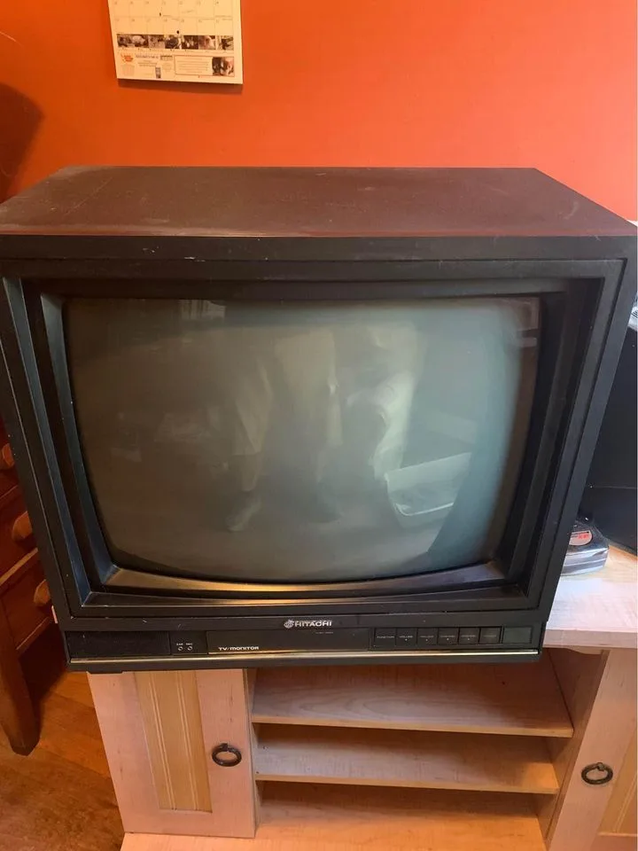 A black Hitachi CRT television sits on a light wood TV stand against an orange wall, its screen dark and reflective, showing…