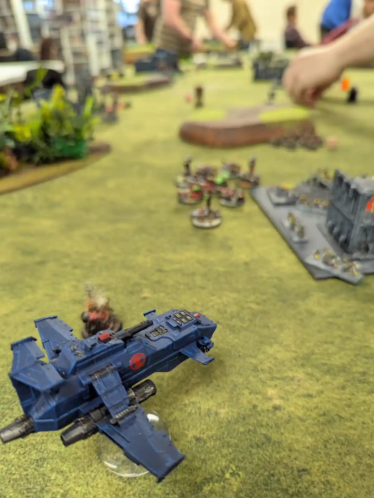 A close-up photo of a blue, winged sci-fi aircraft miniature on a green gaming mat, with other miniature figures and terrain…