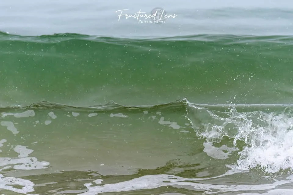 A close-up photograph of a green ocean wave cresting and breaking with white foam, showing water droplets suspended in the…