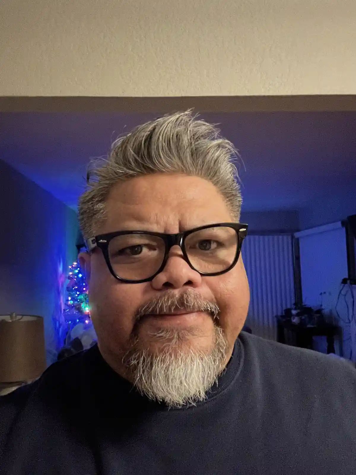 A close-up selfie of a middle-aged man with graying hair and beard, wearing thick black-rimmed glasses and a dark t-shirt…