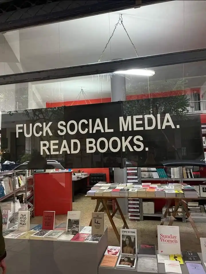 A bookstore window displays a large black banner with bold white text reading 'FUCK SOCIAL MEDIA. READ BOOKS.' Inside…