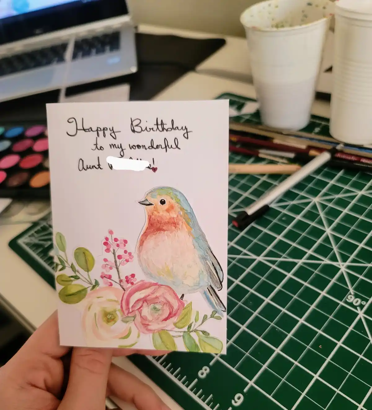 A hand holds a handmade birthday card featuring a watercolor illustration of a bird perched on flowers. The card has…