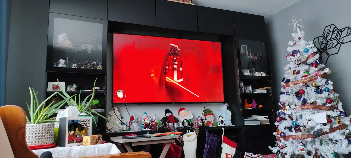 A living room decorated for Christmas with a white tree, stockings, and festive figurines, while a TV displays Darth Vader…