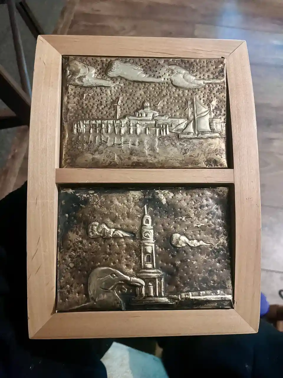 A wooden-framed decorative piece with two metal relief panels. The top panel depicts a coastal scene with a pier, buildings…