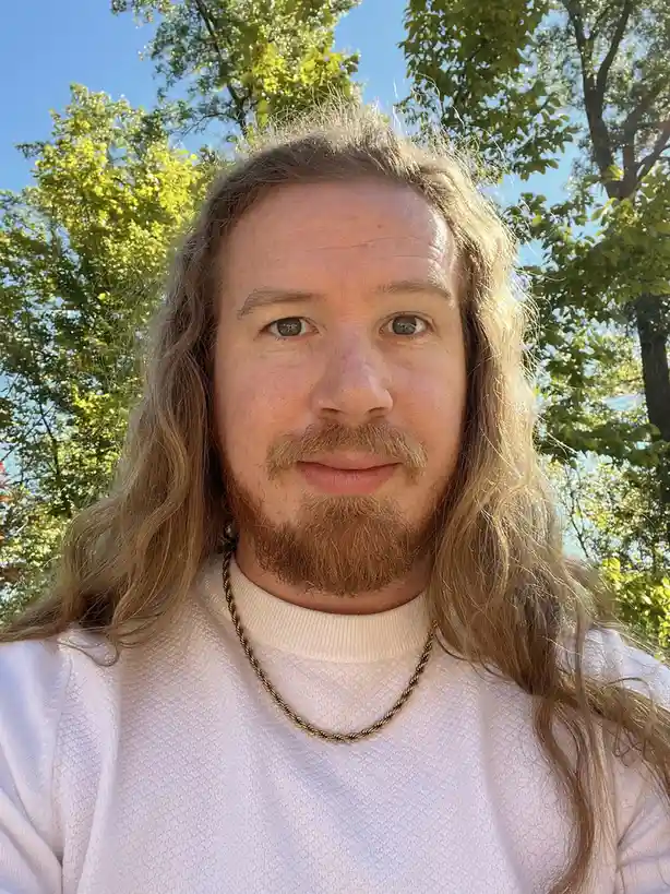 A man with long, wavy, light brown hair and a reddish-brown beard looks directly at the camera with a neutral expression. He…