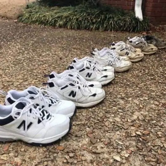 A row of New Balance sneakers laid out on gravel, showing a clear progression from pristine white shoes on the left to…