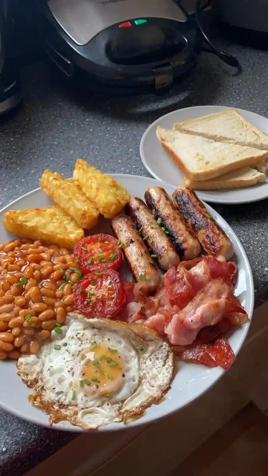 A plate of a full English breakfast featuring fried egg, baked beans, hash browns, grilled sausages, bacon, and halved…