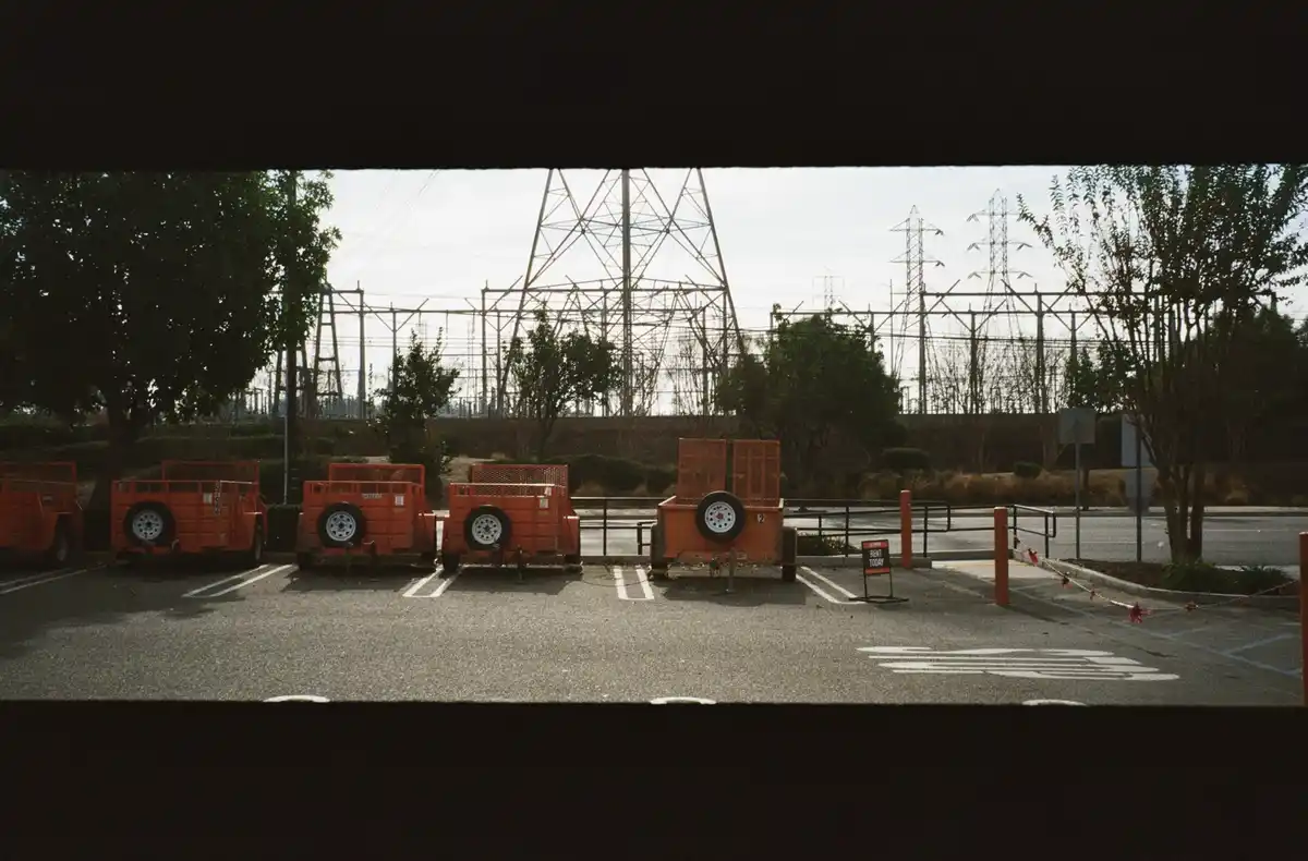 A row of bright orange utility trailers parked in an empty asphalt lot, viewed through a dark horizontal frame. In the…