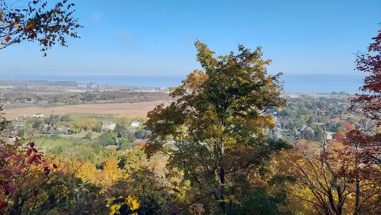 A scenic autumn overlook shows vibrant yellow, orange, and red foliage in the foreground with a sprawling suburban landscape…