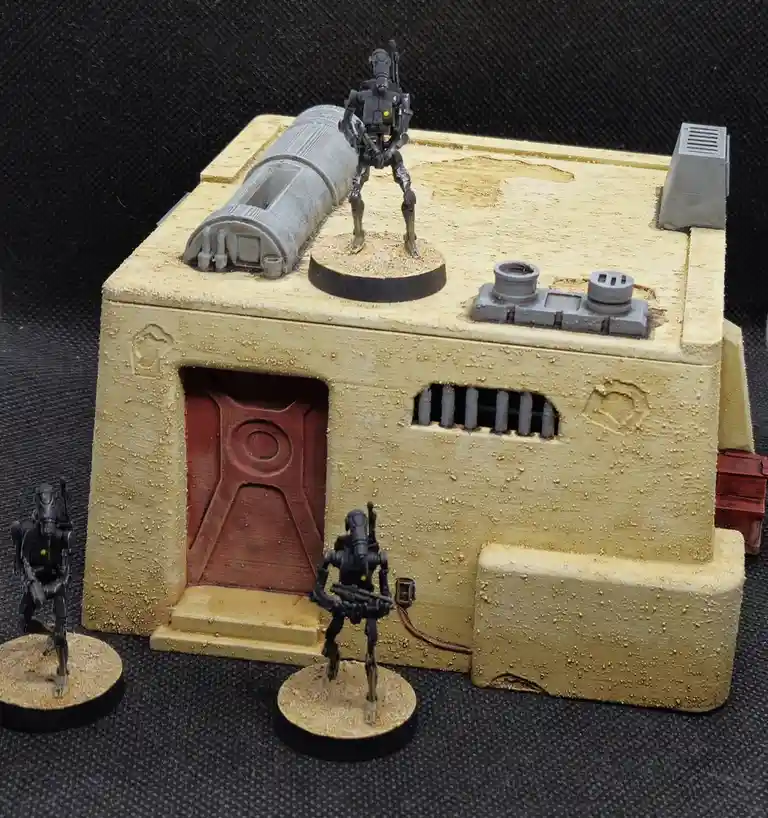 A miniature diorama of a desert-style building with three black, humanoid robot figures. One robot stands atop the…