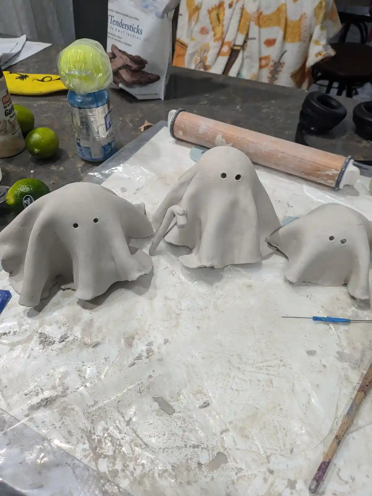 Three handmade clay ghosts with two holes for eyes sit on a messy plastic sheet on a kitchen counter, surrounded by art…