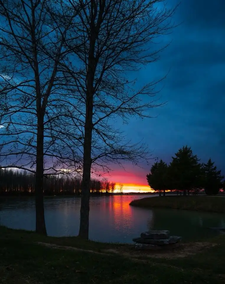 A serene twilight scene at a lake with bare trees silhouetted against a vibrant orange and purple sunset reflected on the…