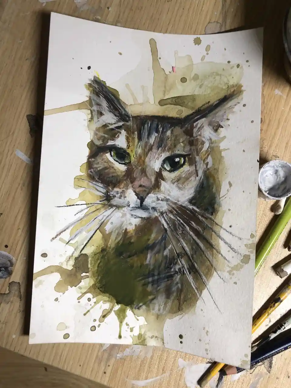A watercolor painting of a cat's face on paper, surrounded by messy paint splatters and drips in brown and green tones, sits…