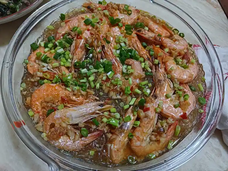 A close-up photo shows a glass bowl filled with steamed shrimp in a savory sauce, topped with chopped green onions and…