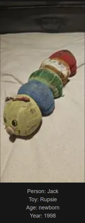 A worn, multi-colored plush caterpillar toy lies on a white fabric surface. The toy has a yellow head with simple eyes and a…