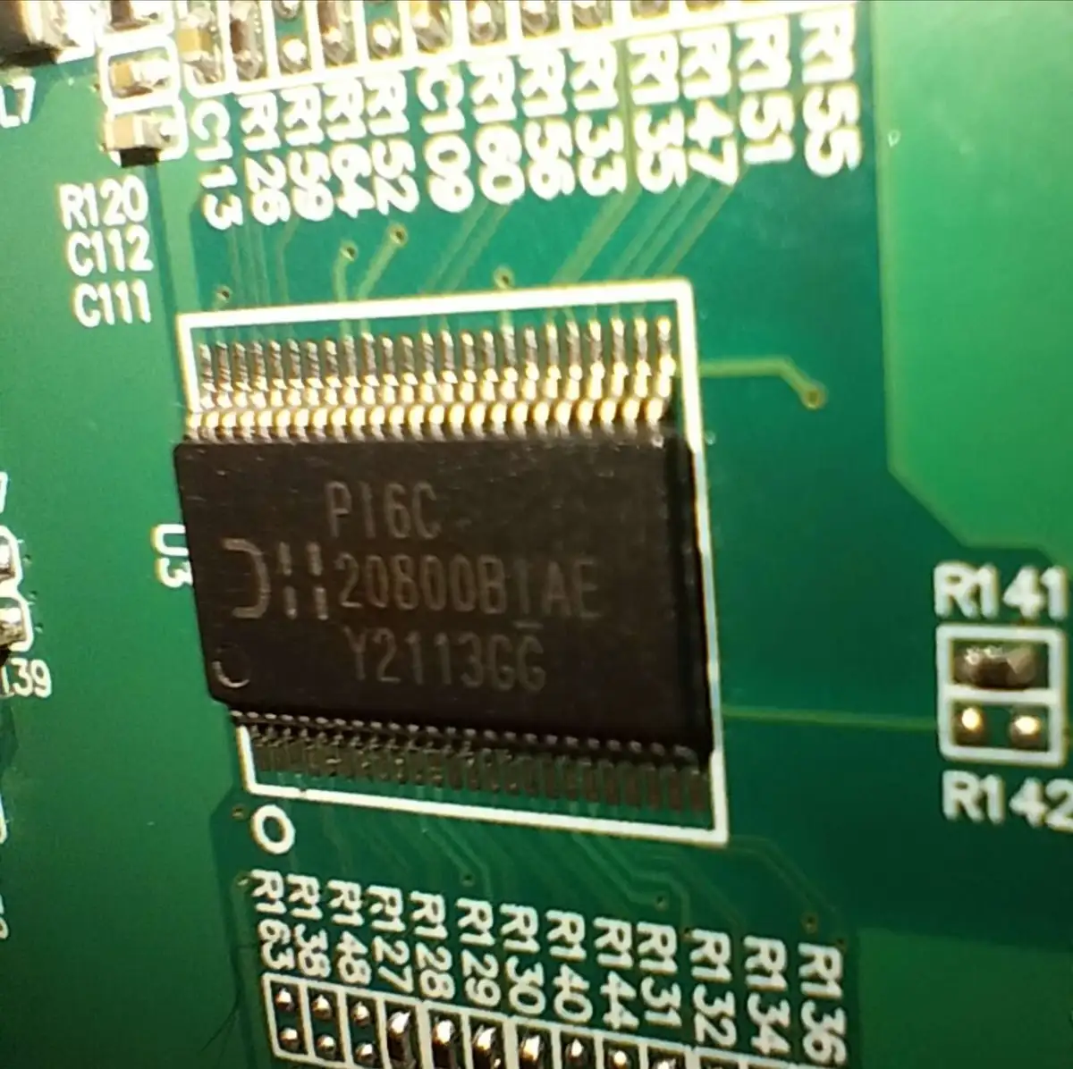 A close-up photo of a black integrated circuit chip mounted on a green circuit board, with visible solder pads and white…