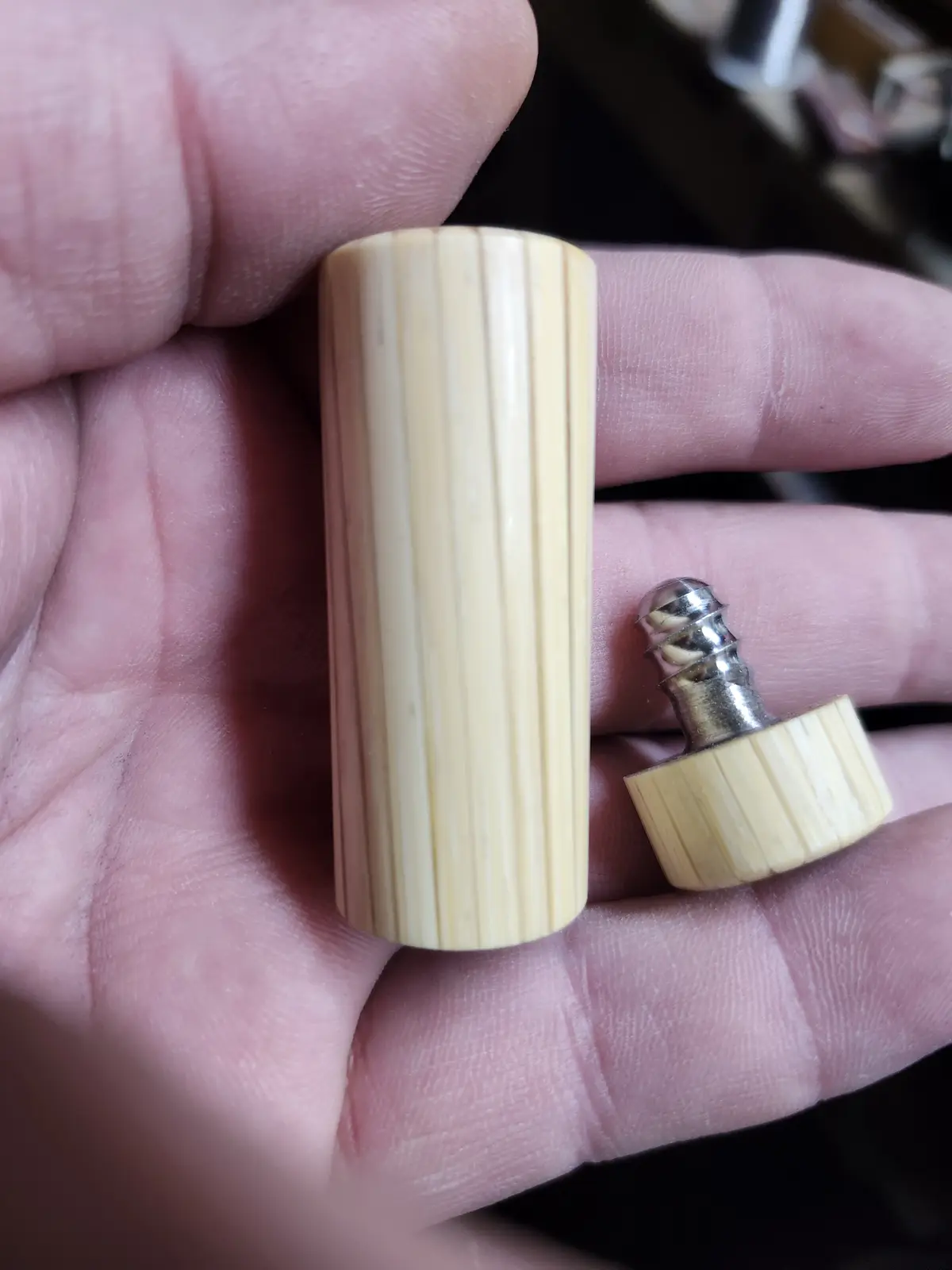 A hand holds a small, light-colored wooden cylinder with vertical grooves, next to its cap which reveals a metal screwdriver…