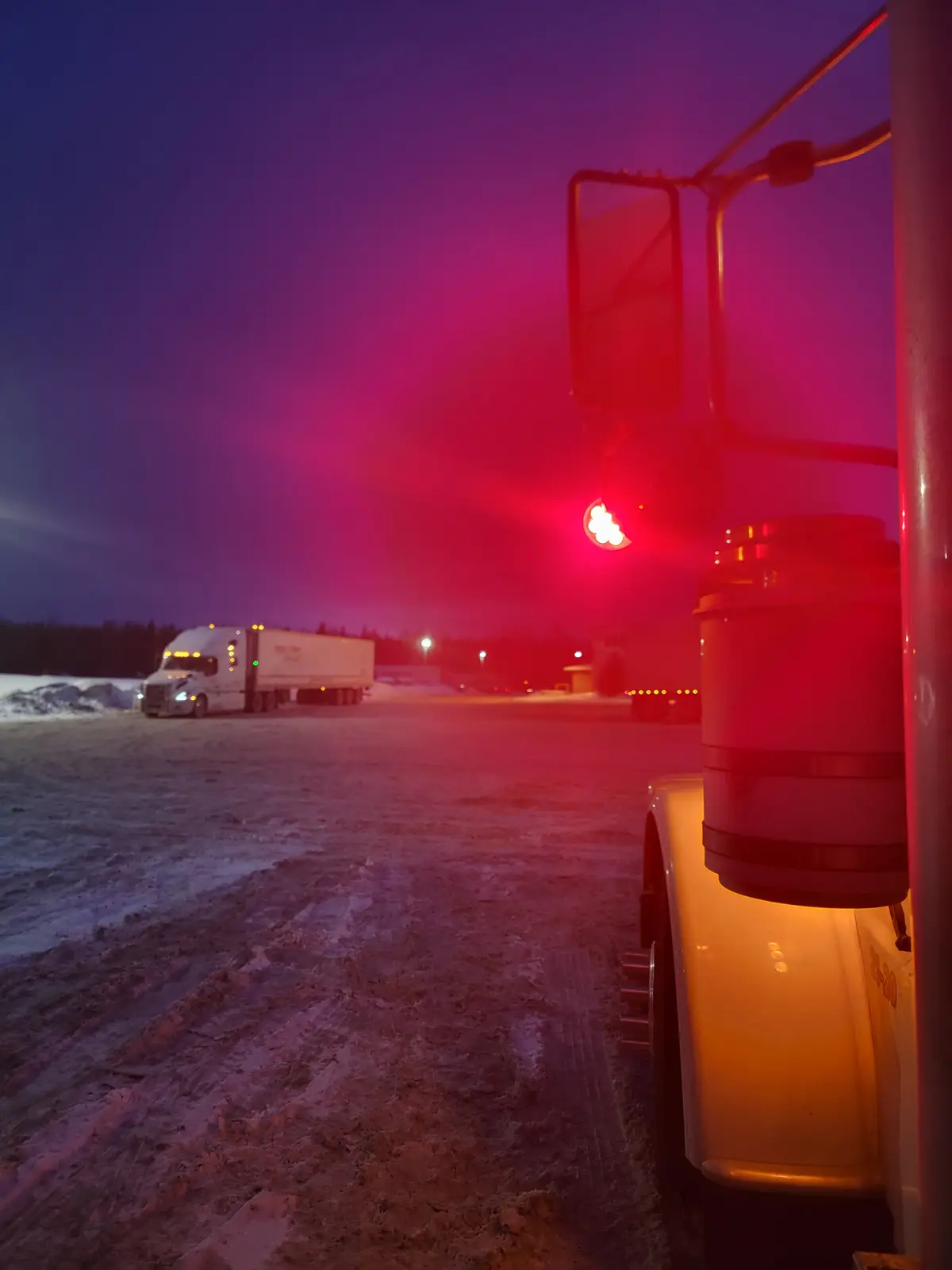 A snowy truck stop at dusk, viewed from the side of a semi-truck with a red light casting a glow. Another semi-truck is…
