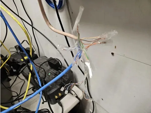 a messy tangle of various colored wires and cables, some stripped and exposed, protruding from a wall outlet or junction…