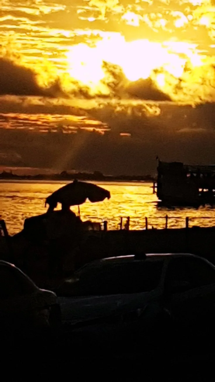 A silhouette of a person standing under an umbrella at a waterfront during a golden sunset, with cars parked in the…