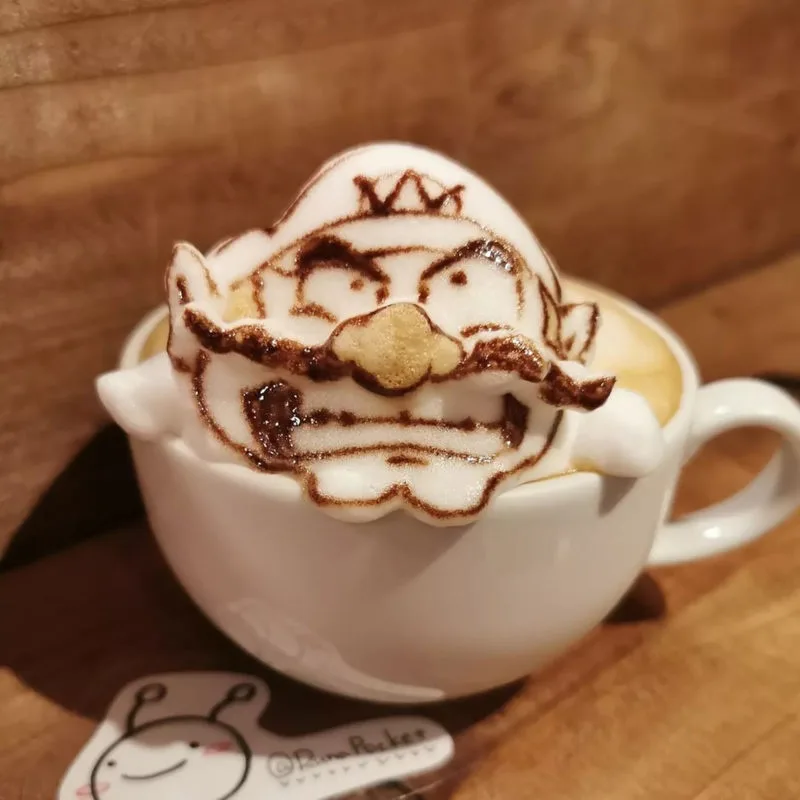 A white ceramic coffee cup filled with latte art depicting the video game character Wario, recognizable by his crown…