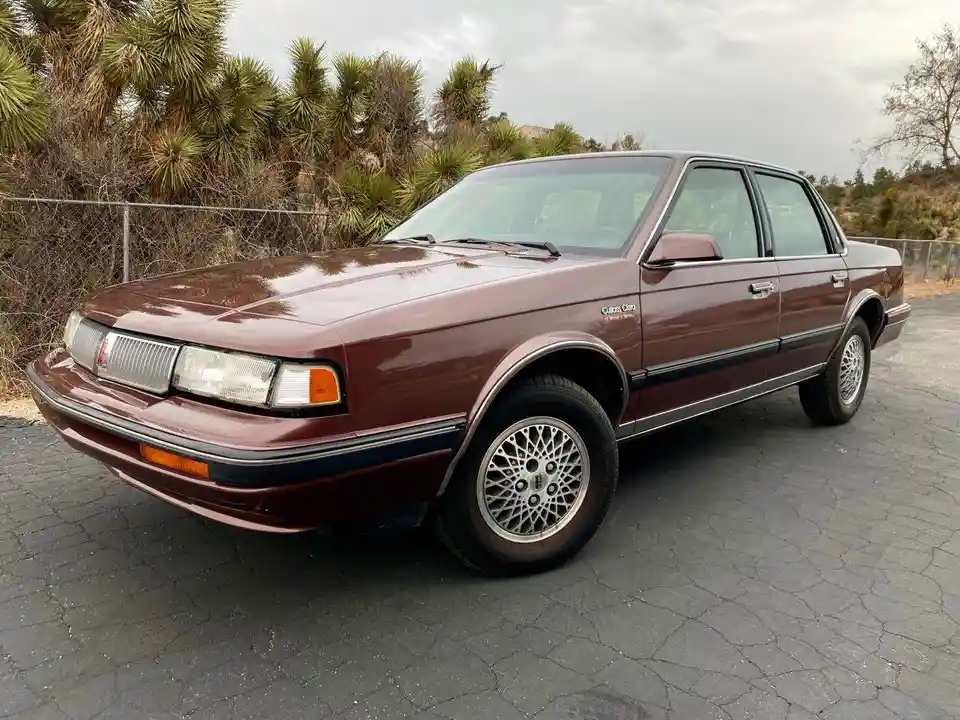 a maroon 1987 buick century sedan is parked on a cracked asphalt surface, with palm trees and a chain-link fence visible in…