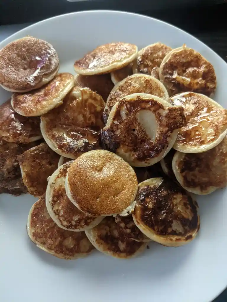 A white plate is piled high with small, golden-brown pancakes, some slightly burnt, drizzled with syrup. The text overlay…