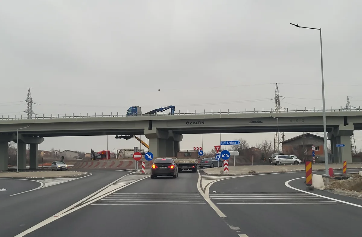 A black car drives on a road under a large concrete bridge with "OZALTIN" and "VISIO" written on its side, while a blue…
