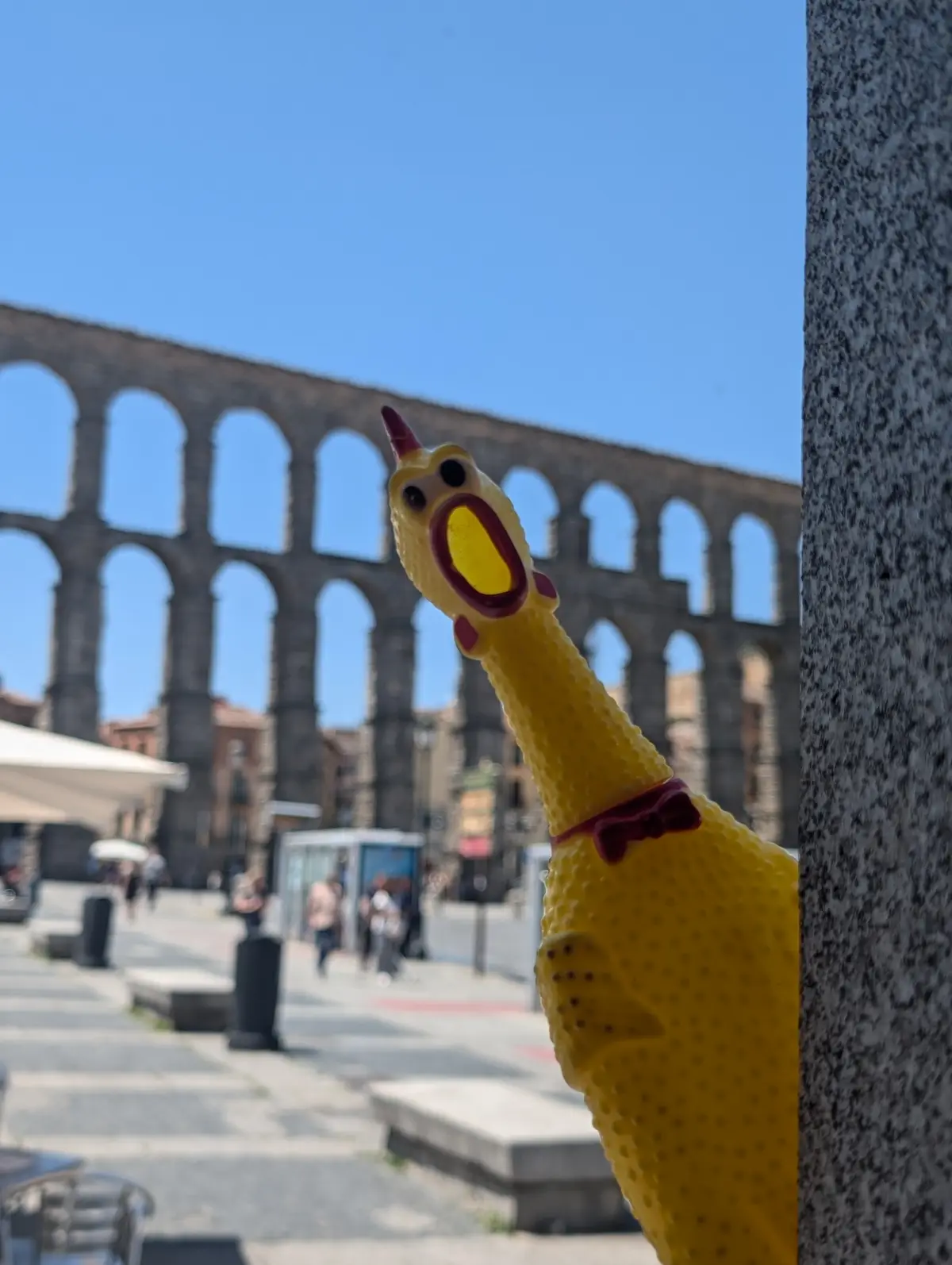A bright yellow rubber chicken toy with a red bowtie and open beak is peeking out from behind a stone pillar, set against a…