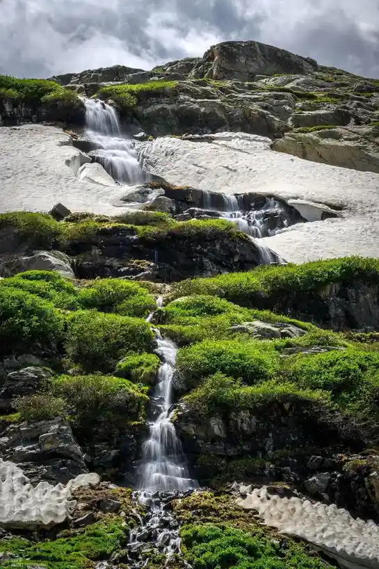 A scenic mountain landscape features a waterfall cascading down rocky terrain interspersed with patches of melting snow and…