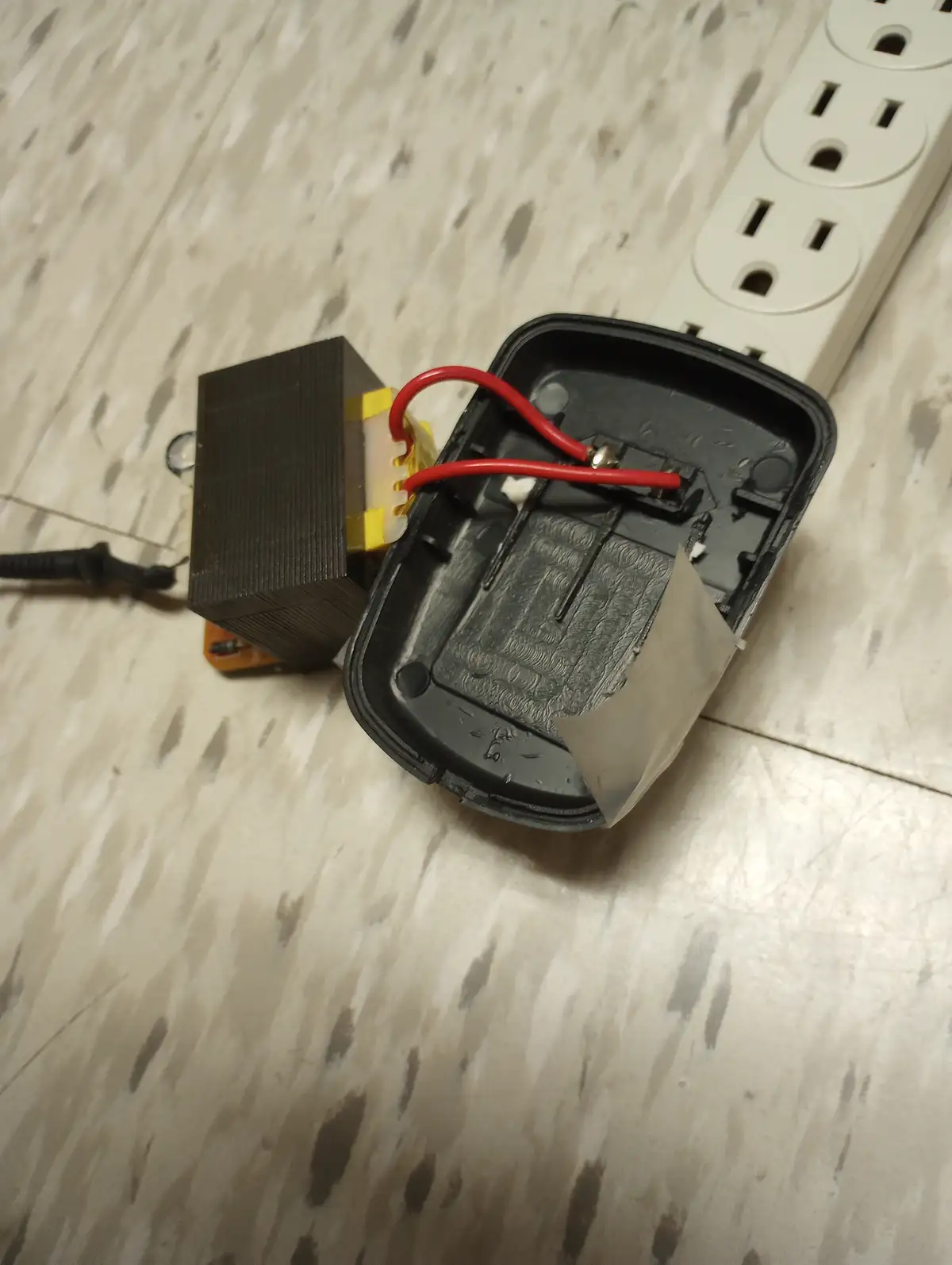 A disassembled black computer mouse lies open on a speckled floor, revealing its internal circuitry and red wires connected…