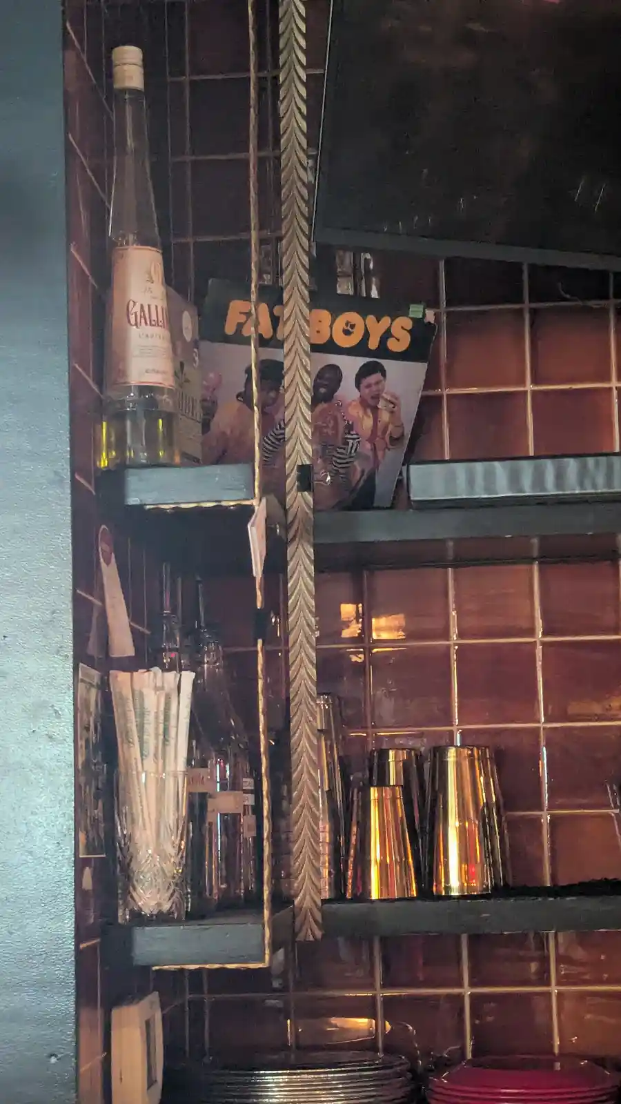 A dimly lit bar shelf with a bottle of Galliano, cocktail shakers, and a poster for the movie 'Fat Boys' visible behind a…