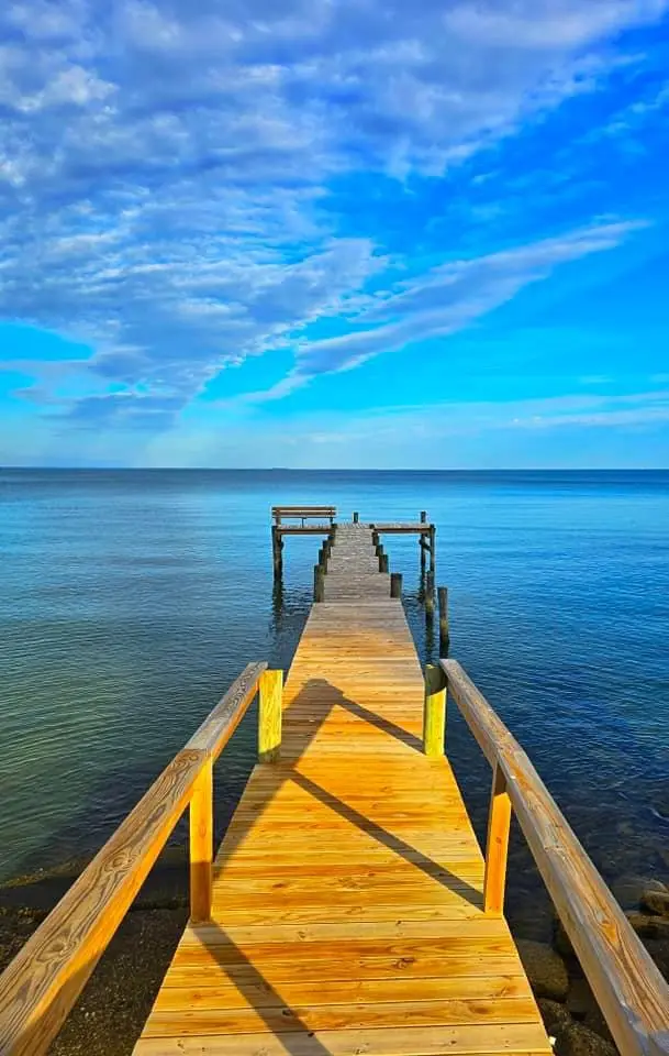 A serene, sunlit wooden pier extends into calm blue water under a vibrant blue sky with scattered clouds. The pier's planks…