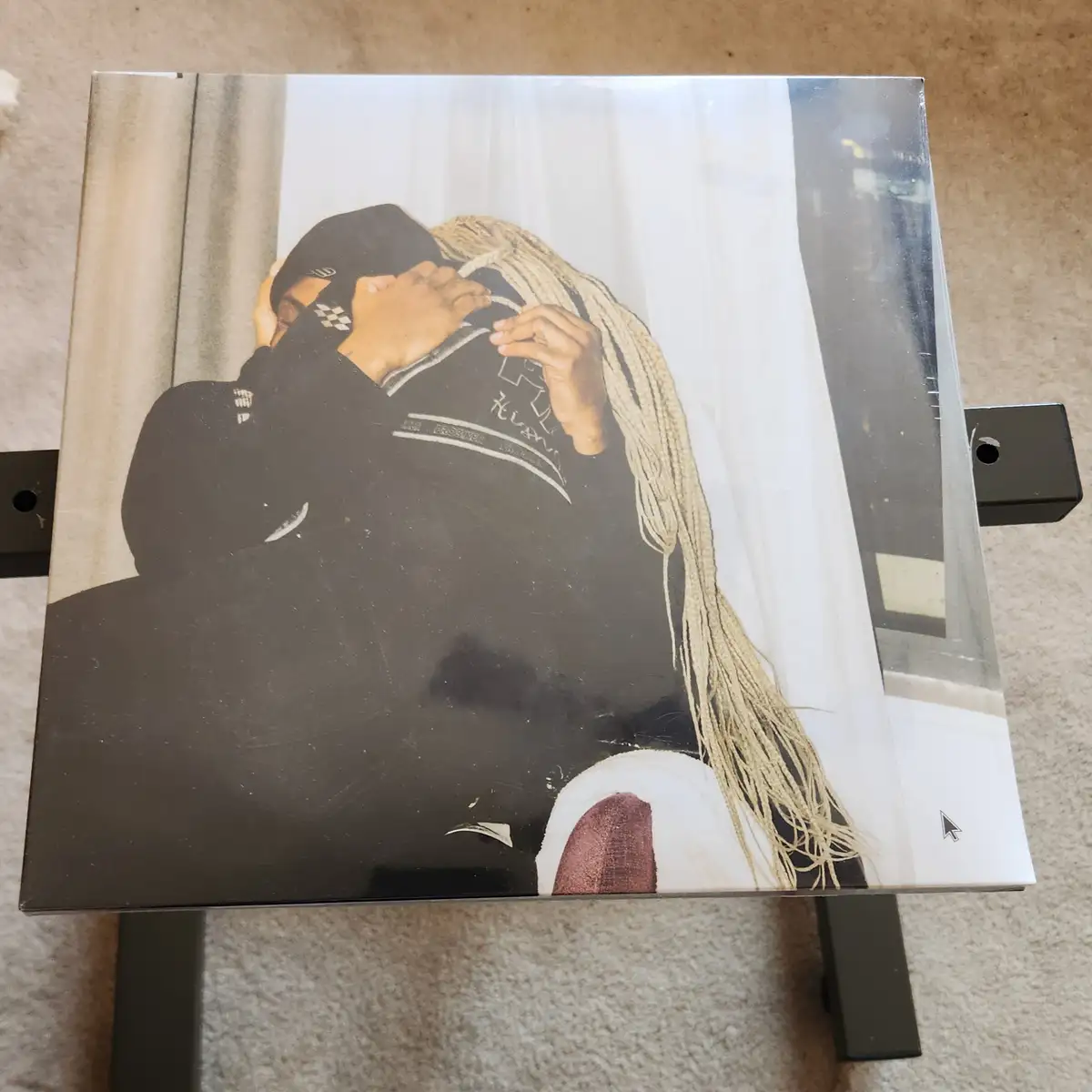 A top-down photo of a sealed album cover featuring a person with long blonde braids wearing a black hoodie and cap, covering…