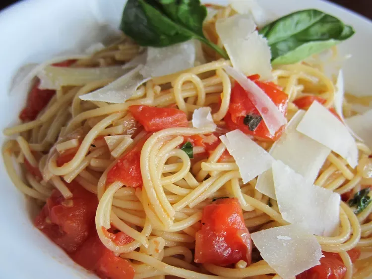 A close-up photo of a white bowl filled with spaghetti tossed in a tomato sauce, garnished with fresh basil leaves and…