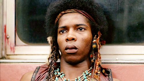 A person with a large afro, braided hair, and colorful beaded necklaces and headwear stares with a deadpan expression. They…
