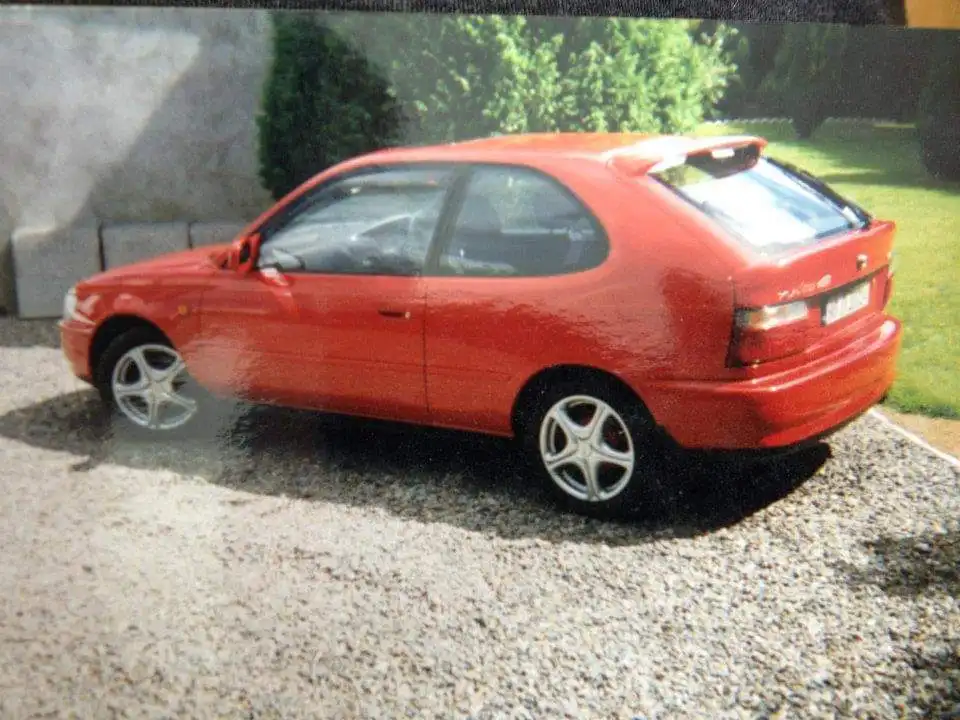 A red Alfa Romeo 147 hatchback is parked on a gravel driveway with green grass and bushes in the background. The car has…