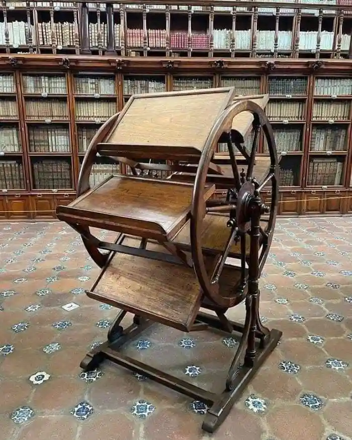 a wooden, antique-style desk with a large wheel mechanism for height adjustment sits in a grand library with tall…