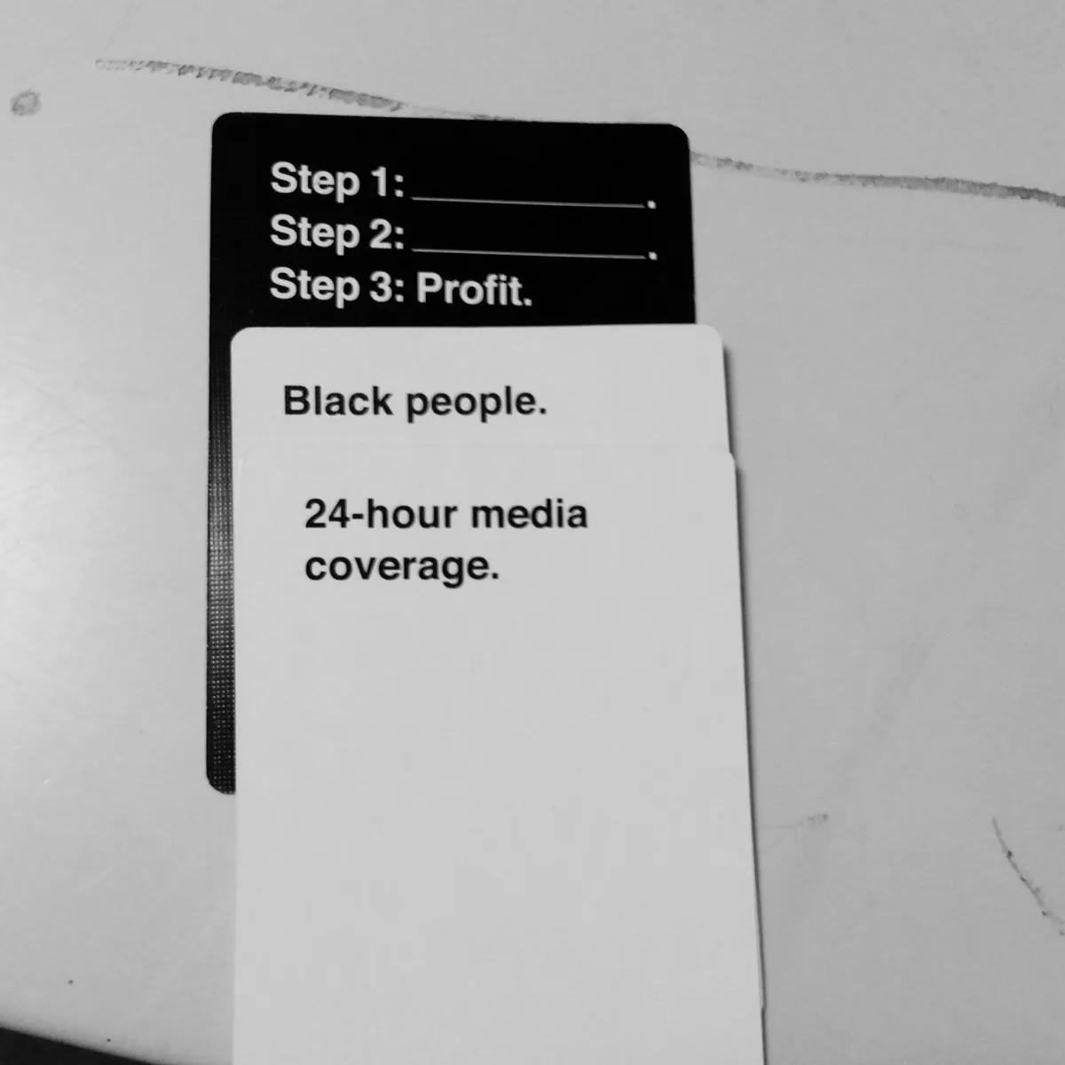 Two cards are stacked on a surface. The top black card reads 'Step 1: ______. Step 2: ______. Step 3: Profit.' The bottom…