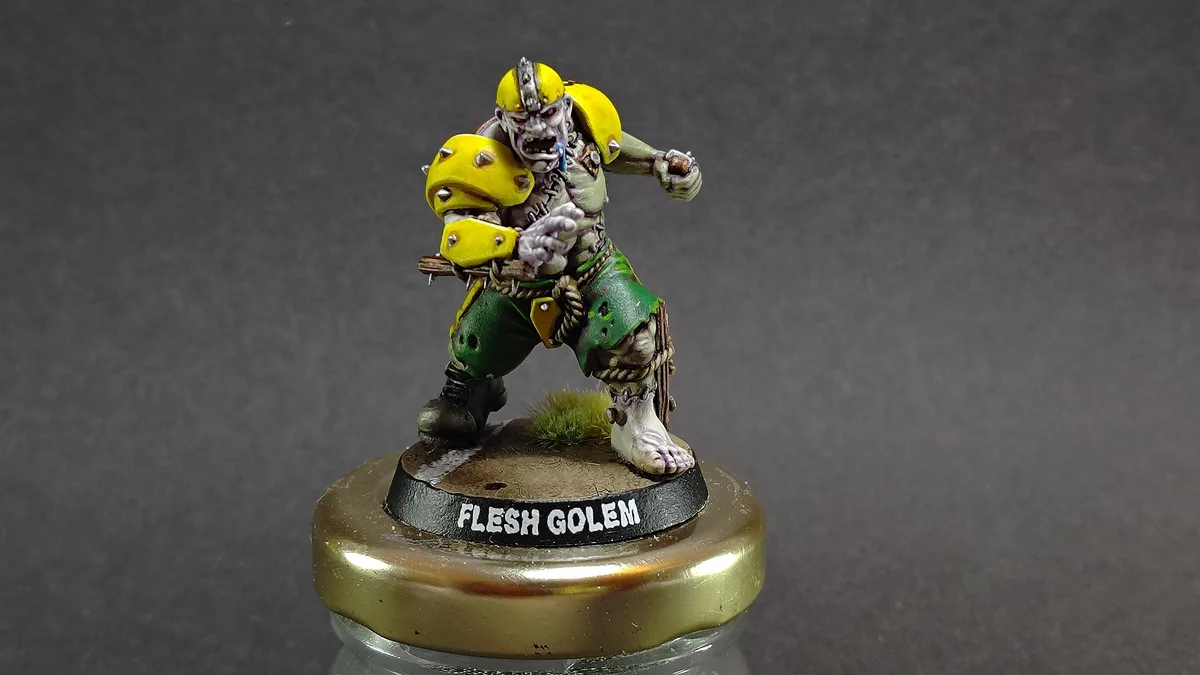 A painted miniature figurine of a grotesque, zombie-like creature with yellow spiked armor and green pants stands on a…
