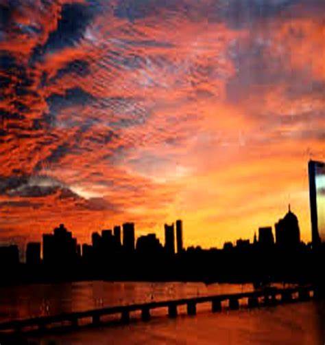 A dramatic sunset with fiery orange and red clouds over a city skyline, silhouetted against the glowing sky. A bridge spans…